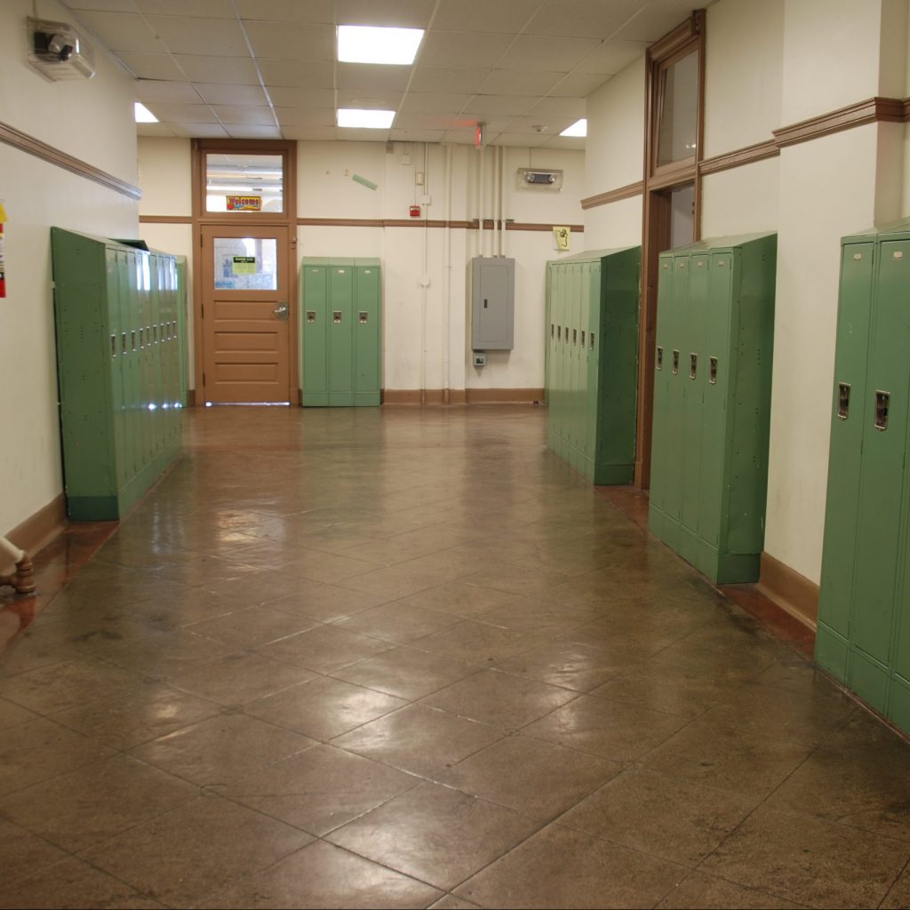 The VCT flooring in this school hallway have recently been cleaned. The green lockers and brown floors along with the off white walls and brown wood trim indicate this is an older school, but the floors have held up well to wear and tear.